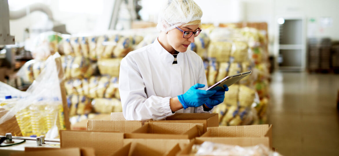 Young beautiful worried female worker in sterile clothes is using a tablet while being leaned against the stacks of open boxes inside of factory storage.